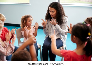 Elevated view of infant school children sitting on chairs in a circle in the classroom, raising hands and learning to count with their female teacher, close up - Shutterstock ID 1279938898