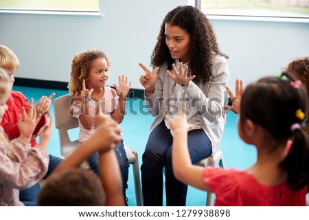 Elevated view of infant school children sitting on chairs in a circle in the classroom, raising hands and learning to count with their female teacher, close up