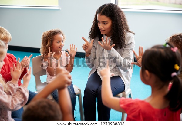 Elevated view of infant school children\
sitting on chairs in a circle in the classroom, raising hands and\
learning to count with their female teacher, close\
up