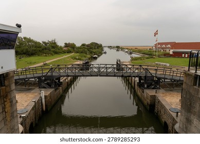 Elevated view from inside a canal lock looking toward a small marina and the open Wadden Sea near Ribe, Denmark. The lock gate and swing bridge span the foreground, framing a calm canal stretching to moored boats, a Danish Dannebrog flag, and a cluster of red and white traditional buildings on the right bank. Green vegetation and flat marshland extend to the horizon under a grey summer sky.
