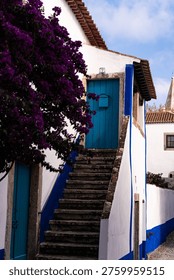 Elevated view looking down a narrow, steep cobblestone street in the medieval fortified town of Óbidos, Portugal. Traditional whitewashed houses with blue and yellow accents and terracotta roofs line the path. The town's iconic church bell tower rises in the distance, set against rolling green hills under a bright sky. People and a few cars are seen on the lower part of the street. A picturesque Mediterranean village scene.