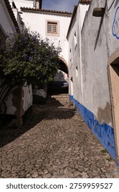 Elevated view looking down a narrow, steep cobblestone street in the medieval fortified town of Óbidos, Portugal. Traditional whitewashed houses with blue and yellow accents and terracotta roofs line the path. The town's iconic church bell tower rises in the distance, set against rolling green hills under a bright sky. People and a few cars are seen on the lower part of the street. A picturesque Mediterranean village scene.