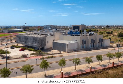 Elevated view of a modern data centre building with extensive rooftop HVAC and cooling tower infrastructure in an urban technology campus near sports facilities and city skyline, Spain