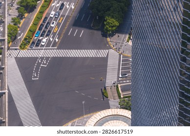 An elevated view over a multi lane downtown junction framed by a glass office tower captures a scooter rider tracing the empty carriageway between crossings. High above the daily commute.