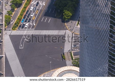 An elevated view over a multi lane downtown junction framed by a glass office tower captures a scooter rider tracing the empty carriageway between crossings. High above the daily commute.
