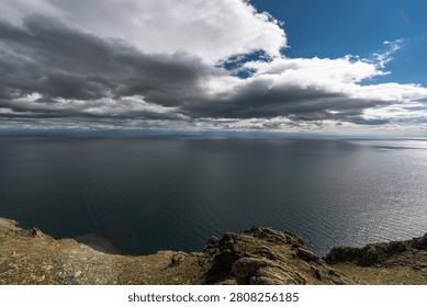 Elevated view from rocky cliffs on Olkhon Island looking out over the vast expanse of Lake Baikal, the world's deepest freshwater lake, in Siberia, Russia. A dramatic contrast of dark storm clouds and bright blue sky dominates the scene, with sunlight breaking through to create a striking silver reflection across the water surface, while a faint rainbow is visible at the cliff base below. Available for editorial and commercial licensing.
