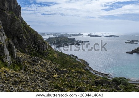 Elevated view shows rugged cliff, clear pale water, rocky islets, and a distant lighthouse on a low island near Henningsvaer, Norway, under soft daylight.
