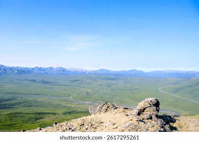 Elevated view of Denali’s vast tundra, winding road, and distant mountain ranges under a clear summer sky from a rocky ridge