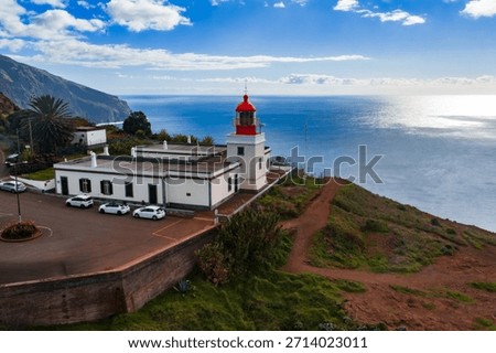 Elevated view of white lighthouse with red lantern at Ponta do Pargo, Madeira, cars in a small lot, red dirt path on a rugged headland, daylight with clouds over the Atlantic.