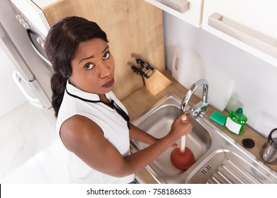 Elevated View Of A Young Unhappy African Woman Using Plunger In Clogged Sink