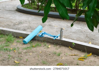 An elevated viewpoint from the front-right captures an uncomplicating outdoor gym layout featuring a blue resistance expander, a rolled mat, and a water flask on a curb.