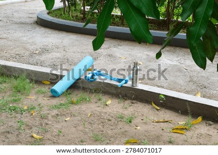 An elevated viewpoint from the front-right captures an uncomplicating outdoor gym layout featuring a blue resistance expander, a rolled mat, and a water flask on a curb.