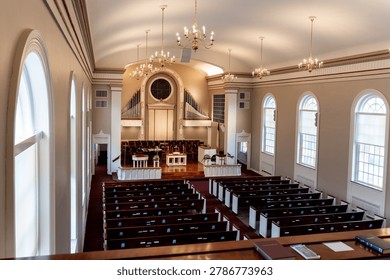 An elevated, wide-angle interior view showcases the traditional sanctuary of Jones Chapel at Meredith College in Raleigh, North Carolina. Row after row of dark wooden pews line the main floor, neatly split by a central carpeted aisle leading toward the altar platform. The front chancel features a large, custom pipe organ integrated into the rear wall beneath a circular window accent. Symmetrical, large arched windows line both sides of the sanctuary, flooding the cream-colored walls with soft, diffused natural light. Elegant brass chandeliers hang from the high, barrel-vaulted ceiling, completing the peaceful and historic architectural setting.