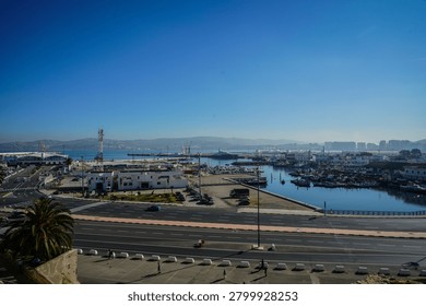 An elevated wide-angle view captures the active Port of Tangier in Tangier, Morocco, during bright daytime. The scene features numerous fishing boats moored in a calm harbor, alongside commercial port facilities, a multi-lane road, and distant mountains under a clear, deep blue sky.