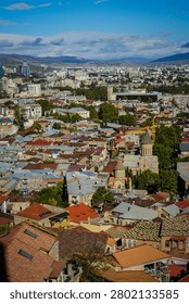 An elevated wide-angle view captures the vibrant urban landscape of Tbilisi, Georgia, with its characteristic mix of ancient and modern architecture. Numerous buildings with colorful tiled roofs and distinctive Georgian Orthodox church domes dot the dense cityscape, extending towards distant hills under a bright clear sky.