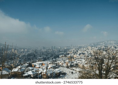 Elevated winter view framed by tree branches, showing dense housing blocks, white rooftops, layered hills and open sky over Sarajevo.