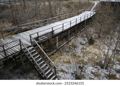 an elevated wooden boardwalk covered in a light dusting of snow, winding through a winter landscape. The pathway, complete with steps and handrails - Powered by Shutterstock