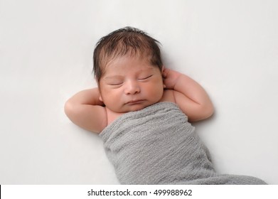 Eleven Day Old Newborn Baby Boy Swaddled In A Gray Wrap. He Is Sleeping On His Back With His Hands Behind His Head On A Cream Colored Blanket.