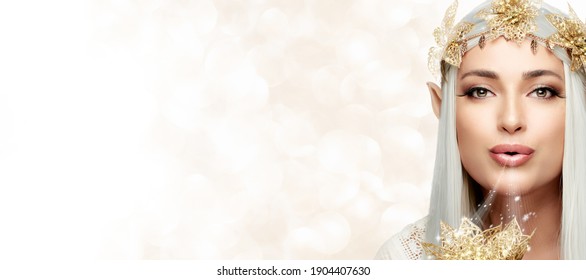 Elf queen with golden flowers wreath looking at camera blowing magic dust. Elves concept banner for carnivals on white and gold background. Beautiful young with elf costume