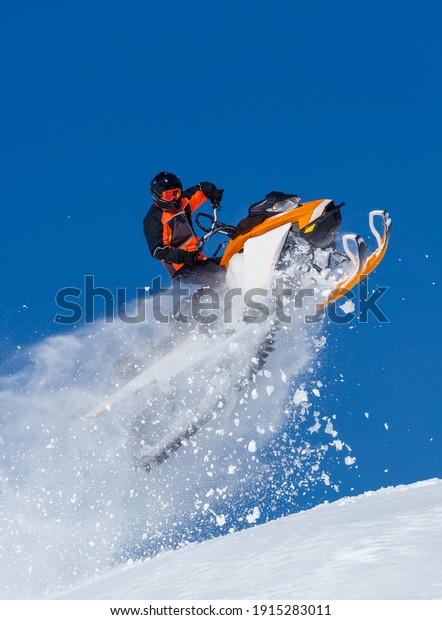 elite sports snowmobiler rides and jumps on steep\
mountain slope with swirls of snow storm. background of blue sky\
leaving a trail of splashes of white snow. bright snowmobile and\
suit without brands