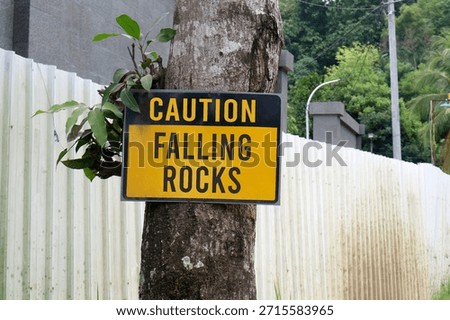 ellow and black Caution Falling Rocks sign mounted on a tree trunk next to a white corrugated fence in a lush green area.