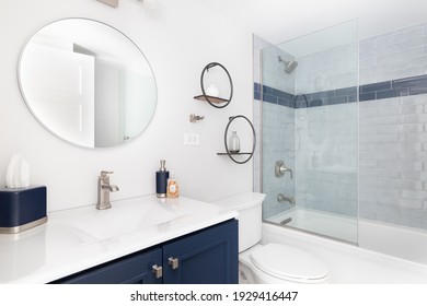 ELMHURST, IL, USA - JANUARY 20, 2021: A Bright White Bathroom With A Blue Vanity Cabinet, White Granite Countertop, Circular Mirror, And A Blue Subway Tile Shower.