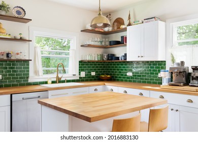ELMHURST, IL, USA - MAY 25, 2021: A renovated kitchen with white cabinets, a natural wood countertop, chairs sitting at the island, a green subway tile backsplash, and a gold pendant light.