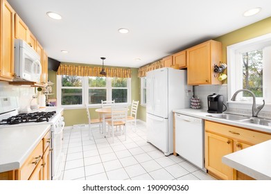 ELMHURST, IL, USA - OCTOBER 2, 2020: A Large Outdated Kitchen With Maple Cabinets, Olive Colored Walls, White Appliances, And A Small Dining Room Table Under A Hanging Light.