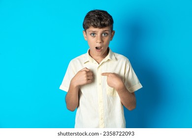 Embarrassed beautiful kid boy wearing casual T-shirt indicates at herself with puzzled expression, being shocked to be chosen to participate in competition, hesitates about something
