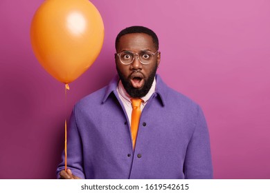 Embarrassed Black Man Stares At Camera, Feels Stunned And Shocked, Comes On Stag Party, Going To Congratulate Best Friend, Holds Balloon, Wears Festive Clothing, Isolated Over Bright Lilac Background