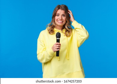 Embarrassed Girl Holding Microphone Awkward Smiling And Scratch Head As Singing Song On Karaoke With Friends, Shy Perform In Front Of Audience, Standing Blue Background