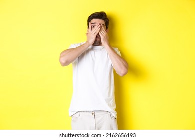 Embarrassed Guy Shut Eyes But Peeking Through Fingers At Something Awkward, Standing Over Yellow Background