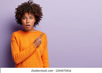 Embarrassed impressed black ethnic woman points fore finger at upper right corner, thrilled by something, shows blank space, wears orange clothing, stands over purple background. Wow, look there