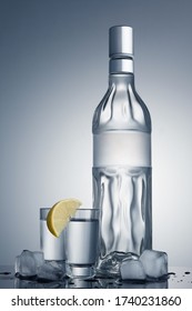 Embossed Vodka Bottle With An Empty Label With Space For Text And Logo. Mockup Of A Transparent Alcohol Bottle On A Gray Gradient Background With Two Glasses, A Slice Of Lemon And Ice