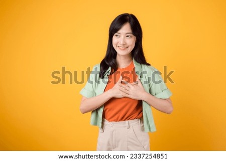 Embrace the love of deals with a young Asian woman 30s wearing a green shirt on an orange shirt, radiating happiness. Love deal promotion concept against a vibrant yellow backdrop.