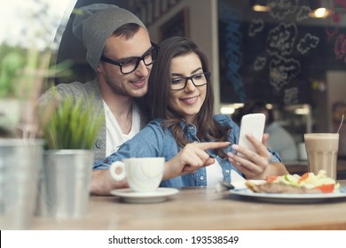 Embracing Couple Using Mobile Phone In Cafe 