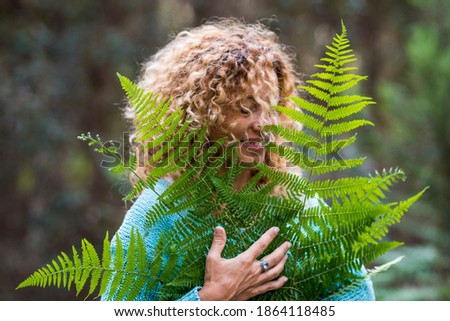 Embracing outdoors concept and love nature with portrait of beautiful young adult woman with big forest leaf and green wood in background - earth's day and care of outdoor nature