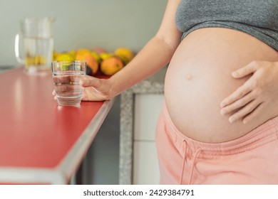 Embracing the vital benefits of water during pregnancy, a pregnant woman stands in the kitchen with a glass, highlighting hydration's crucial role in maternal well-being - Powered by Shutterstock