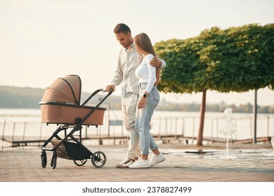 Embracing the woman by the waist. A young couple with a baby pram is walking together.