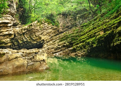 An emerald pool rests beneath tilted limestone layers and mossy cliffs in a Georgian forest gorge, conveying a peaceful and secluded wilderness atmosphere