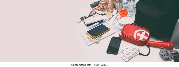 Emergency backpack equipment organized on the table. Documents, water,food, first aid kit and another items needed to survive. - Shutterstock ID 2136493391