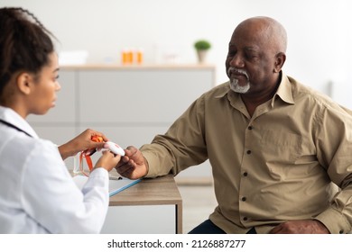 Emergency Button Concept. Portrait Of Black Medical Worker Holding Personal Alarm Button And Giving It To Senior Patient, Sitting At Table In Office. Older Male Looking At Medical Alert