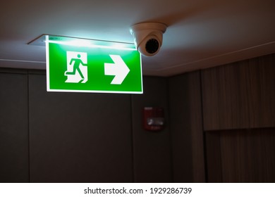 Emergency exit sign at the corridor in building. Green fire exit sign hanging on ceiling on dark corridor in building near fire emergency exit door. Green emergency exit sign. 