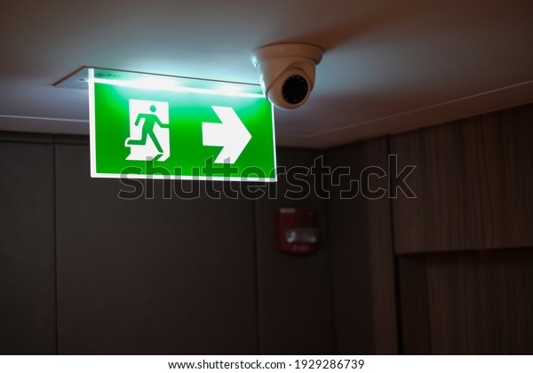Emergency exit sign at\
the corridor in building. Green fire exit sign hanging on ceiling\
on dark corridor in building near fire emergency exit door. Green\
emergency exit sign. 