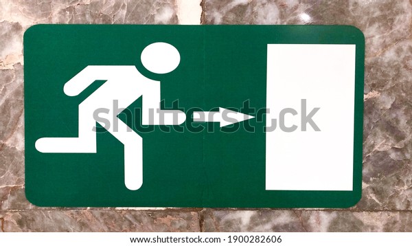 Emergency Exit\
sign on airport. Illuminated green exit sign on the wall in a\
public transportation facility. Signage consists of a human figure\
running and an arrow pointing at a\
door.