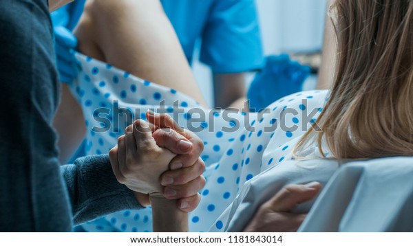 Emergency In the Hospital: Woman\
Giving Birth, Husband Holds Her Hand in Support, Obstetricians\
Assisting. Modern Delivery Ward with Professional\
Midwives.