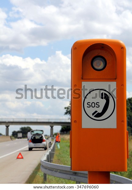 Emergency telephone at the\
roadside\
Emergency telephone on a German highway (Autobahn) with\
car broken down in the background and driver waiting behind crash\
barrier