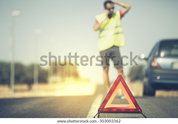 Emergency triangle on the road and man\
with reflective vest calling by phone to car\
assistance