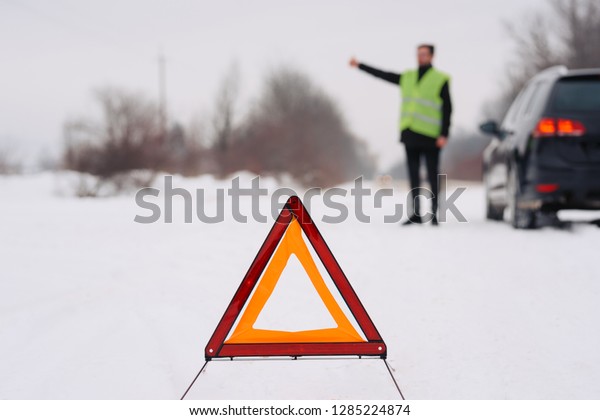 Emergency triangle on the road. Stopped car\
hand up in the\
background.