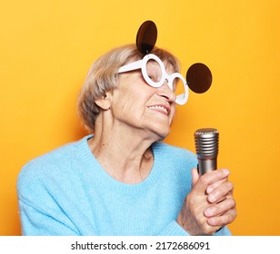 Emotion, Modern Lifestyle And Old People Concept: Happy Old Woman Singing With Microphone Over Yellow Color Background, Having Fun